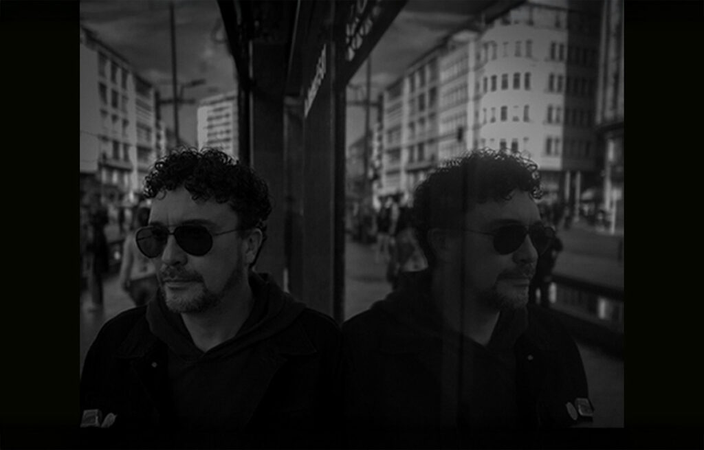 A man with curly hair and sunglasses stands next to a reflective surface in a cityscape. His reflection mirrors him. The scene is monochromatic, creating a contemplative mood.