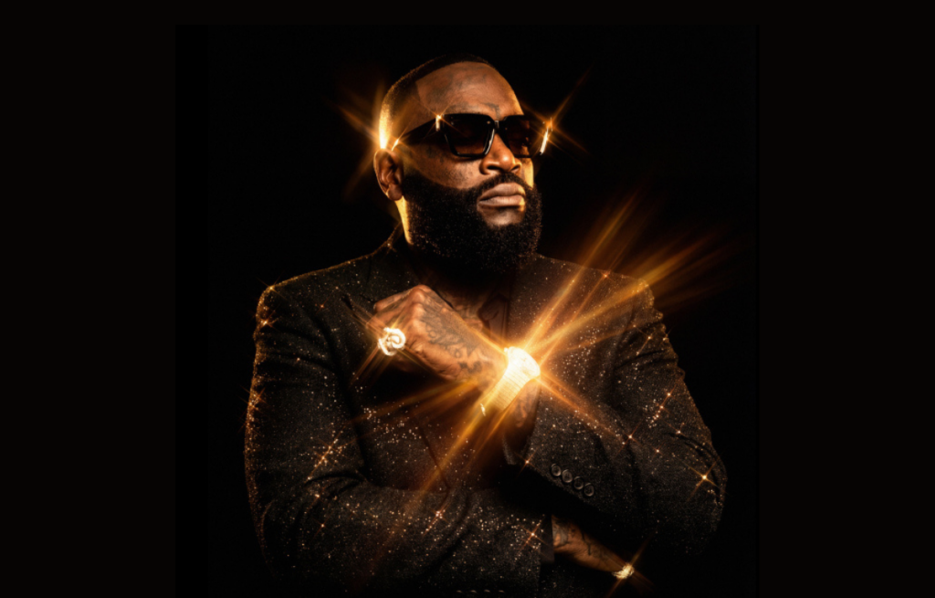 Man in a sparkling black suit and sunglasses stands confidently with crossed arms. Bright golden light radiates from his jewelry, creating a dramatic effect.